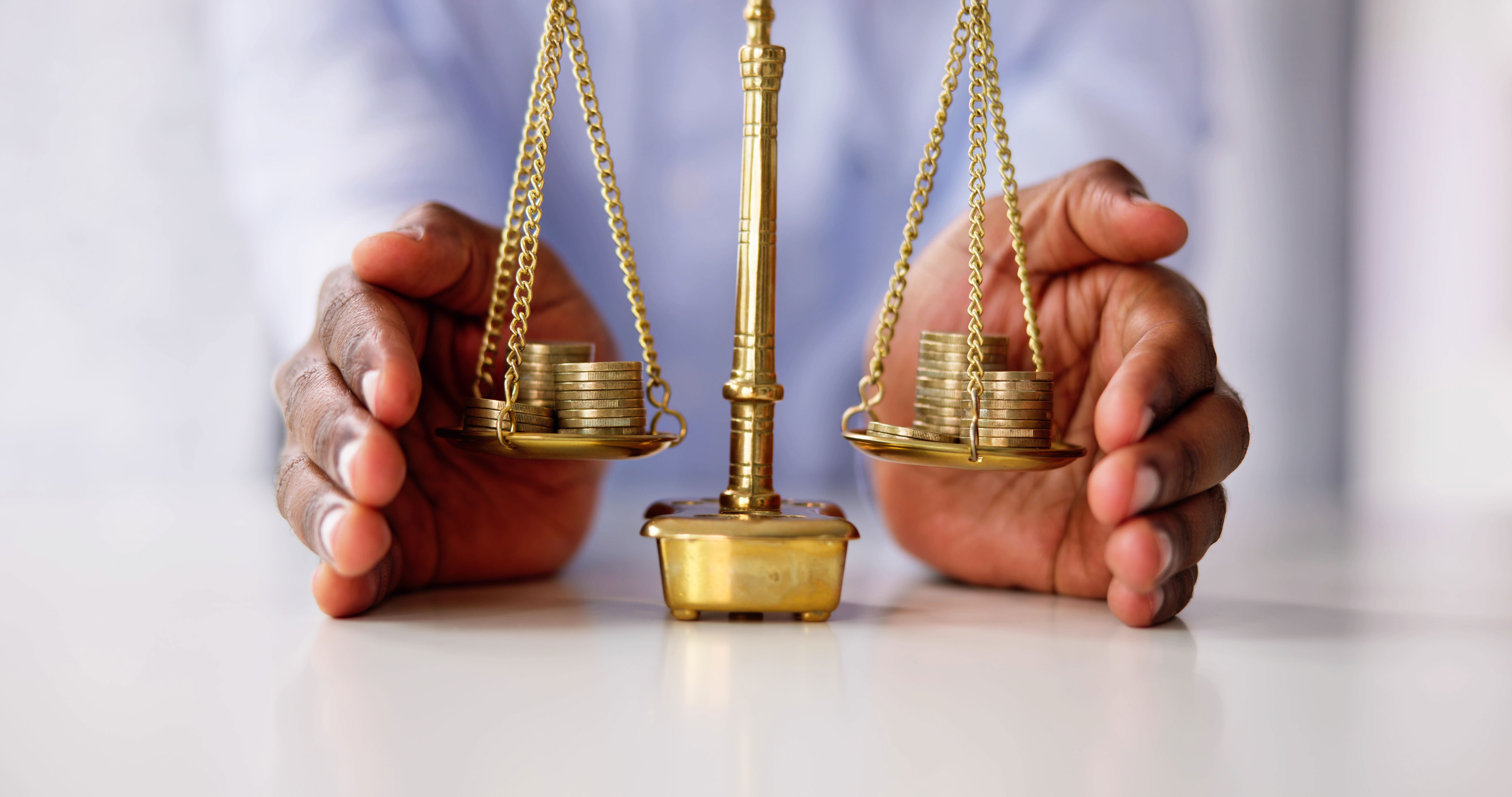 A person holds a balanced brass scale with stacks of coins on both sides, placed on a white surface.